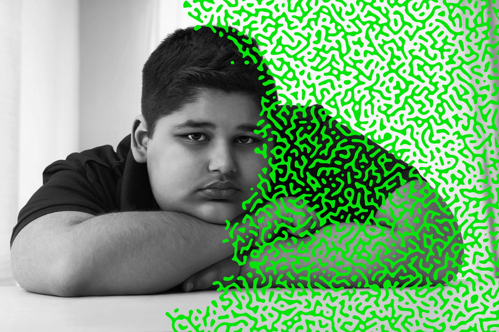 A latino teen boy looks at the camera with his head resting on his folded arms with a bright green overlay of an organic brain-like pattern covering the left half of the image